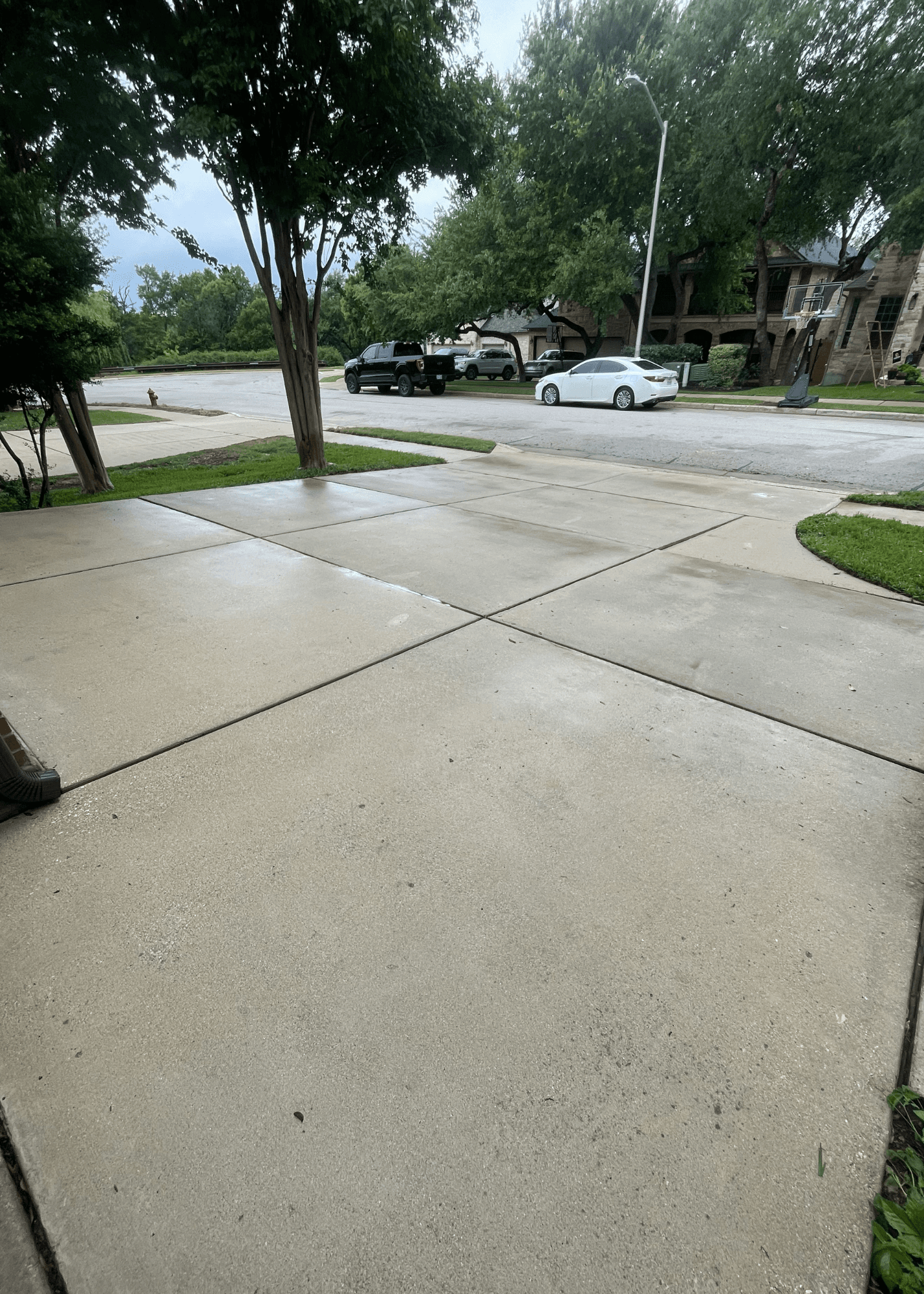 Clean concrete driveway in a suburban neighborhood with parked cars and lush green trees.