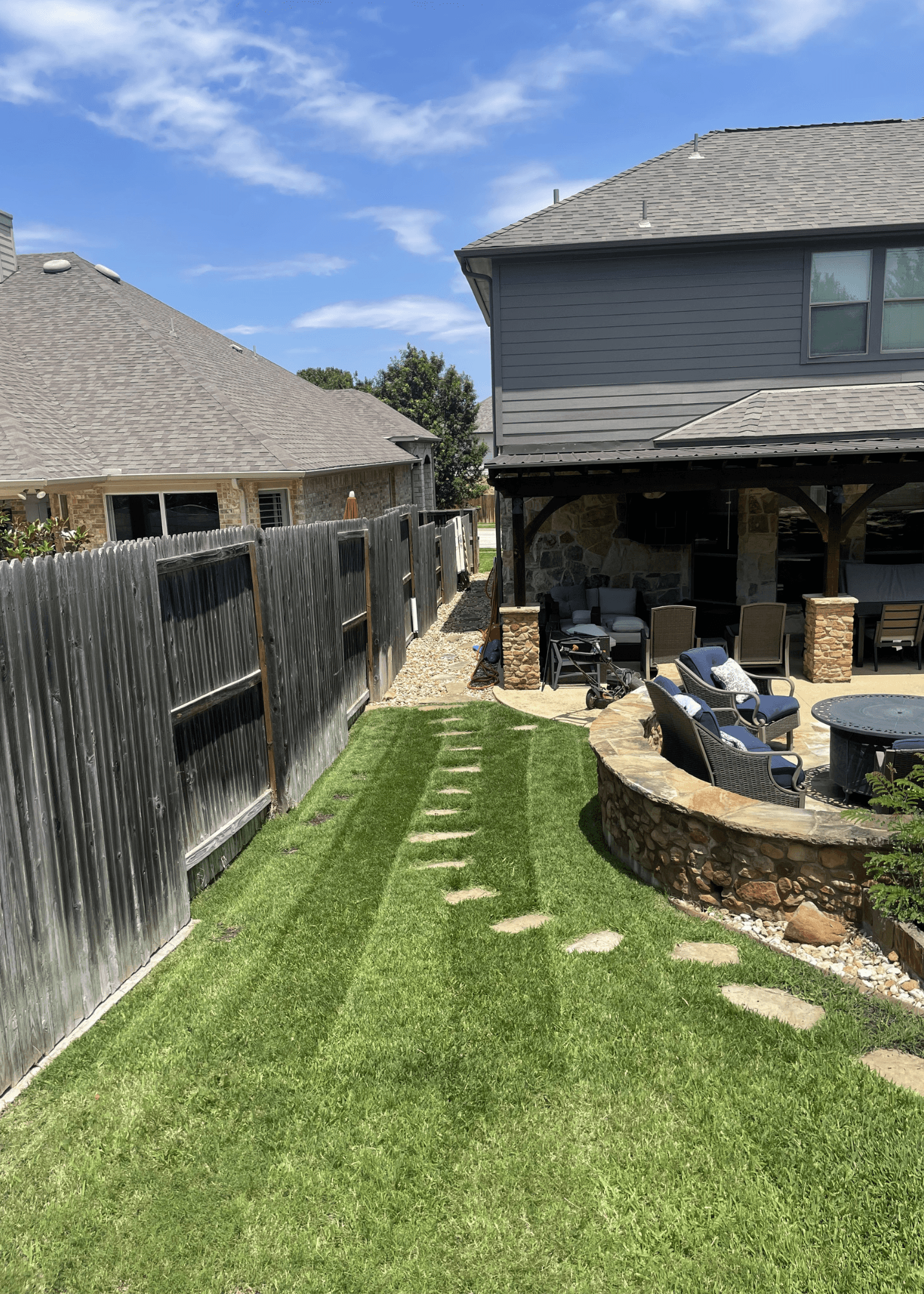 Stepping stones on a green lawn lead past a stone wall to a furnished patio.